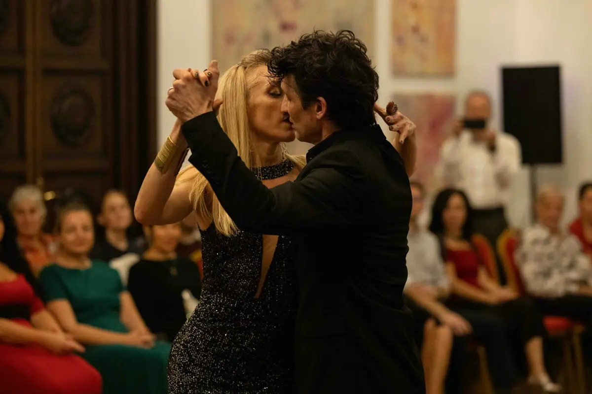 A tango couple in close embrace dancing in front of seated guests, with the woman's eyes closed