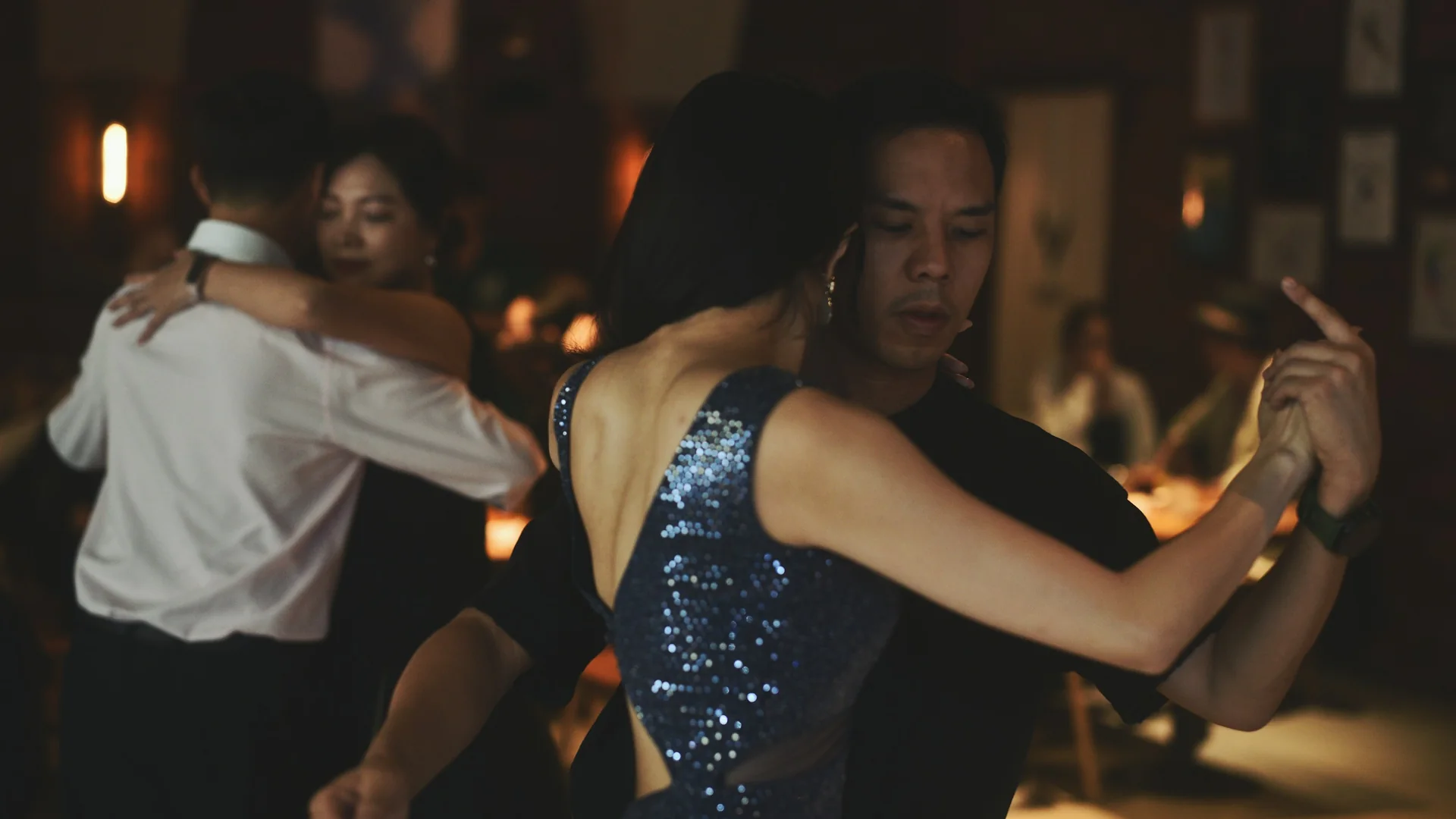 Et tangopar i tæt omfavnelse på et svagt oplyst dansegulv, kvinden i en glitrende blå kjole, med endnu et par dansende bag dem